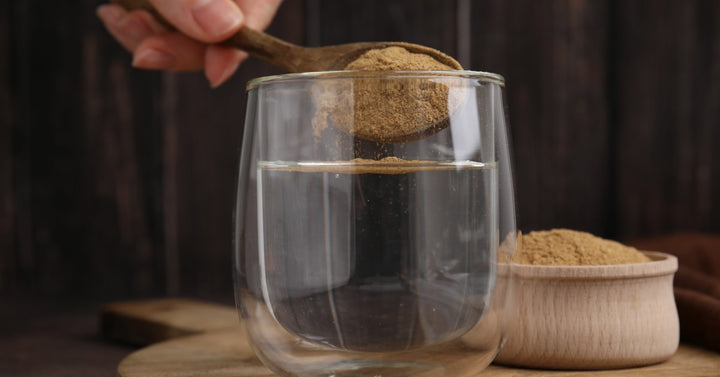 A person's hand takes a wooden spoon and drops a spoonful of powdered supplement into a glass of water.