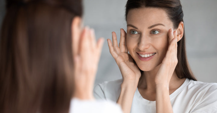 A woman looks at her face in a mirror while touching her cheeks. She has brown hair and wears a white shirt.