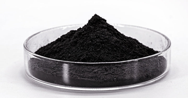 A mound of black powder sits in a transparent, glass petri dish. It sits against a solid white background.